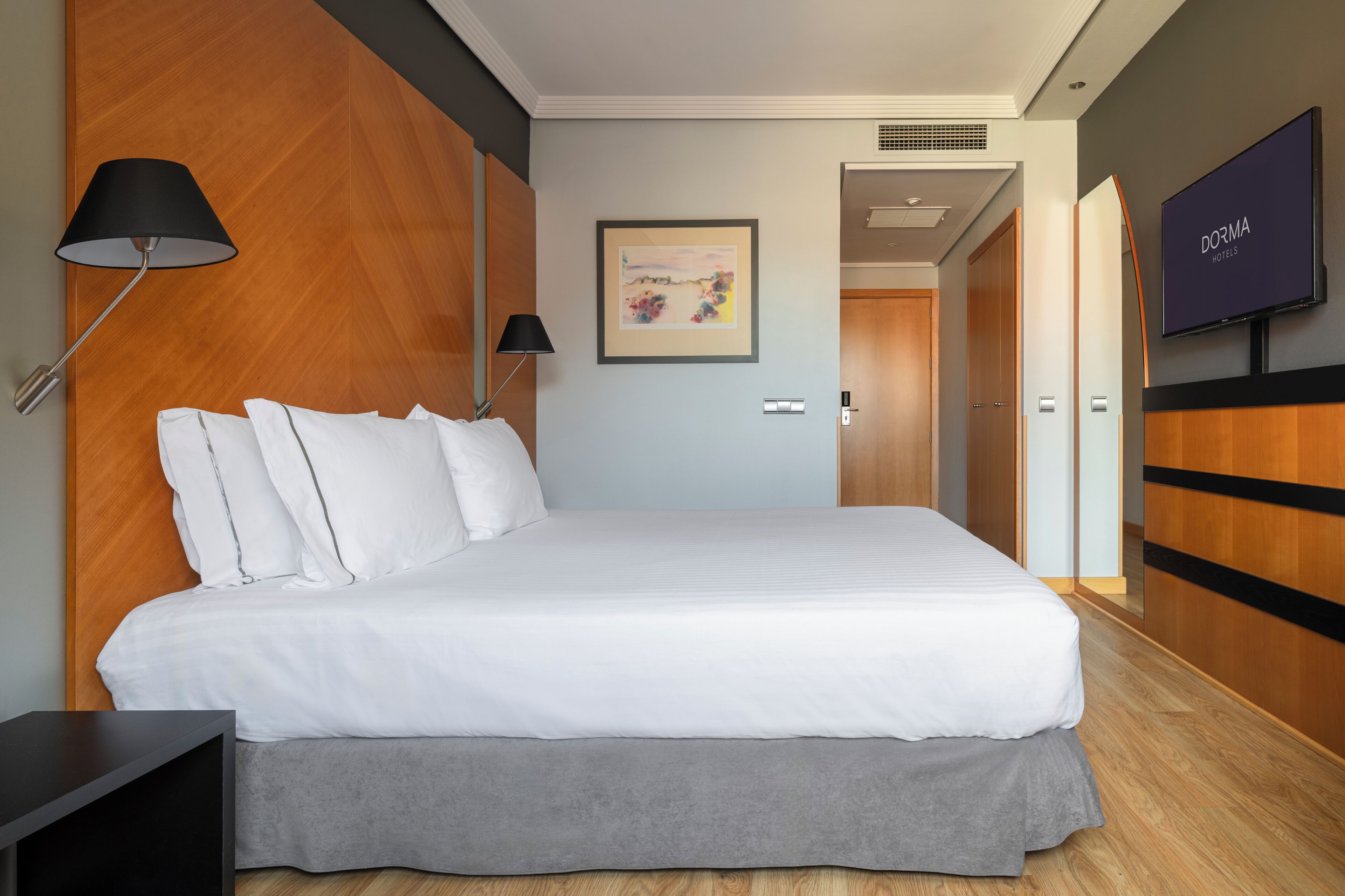 A modern hotel room with a large bed, a TV, a framed picture on the wall, and a wooden wardrobe.