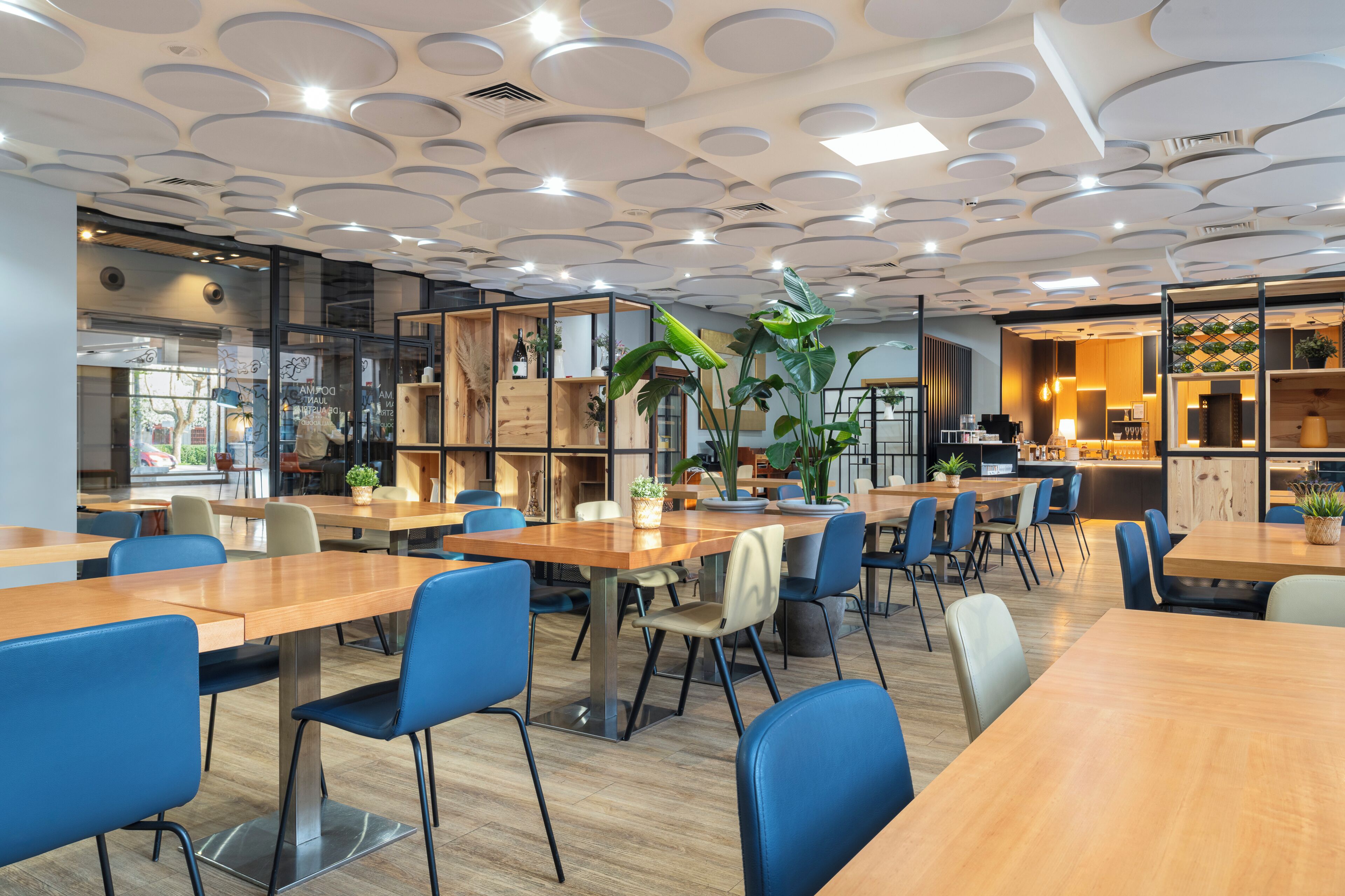 Een moderne eetruimte met houten tafels en blauwe stoelen, een keuken met houten kasten, en een plafond met ronde lampen.