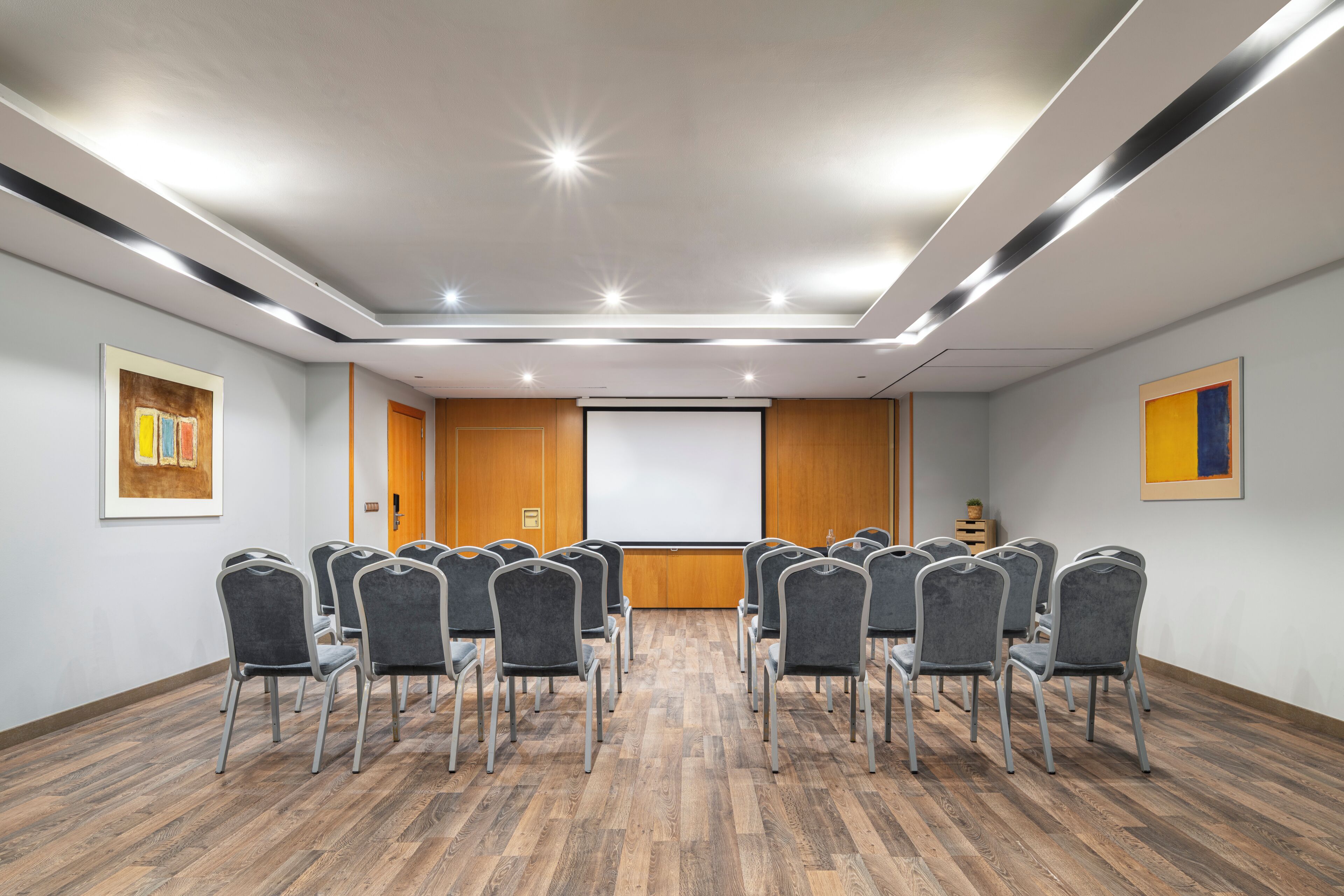 Un salón de conferencias con una pantalla de proyector, filas de sillas y piso de madera.