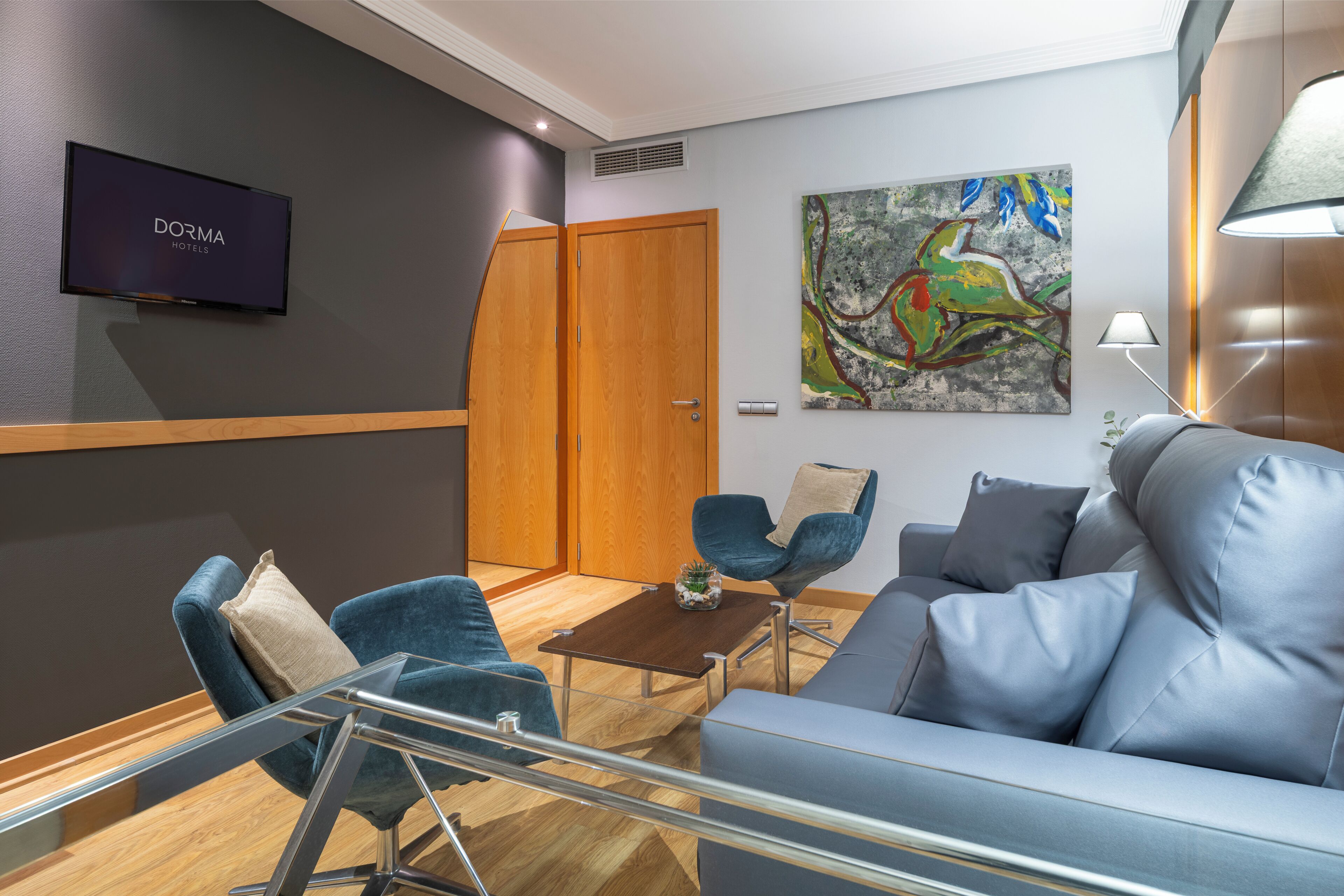 Una habitación de hotel moderna con televisor de pantalla plana, un sofá, dos sillones, una mesa de centro y un cuadro en la pared.