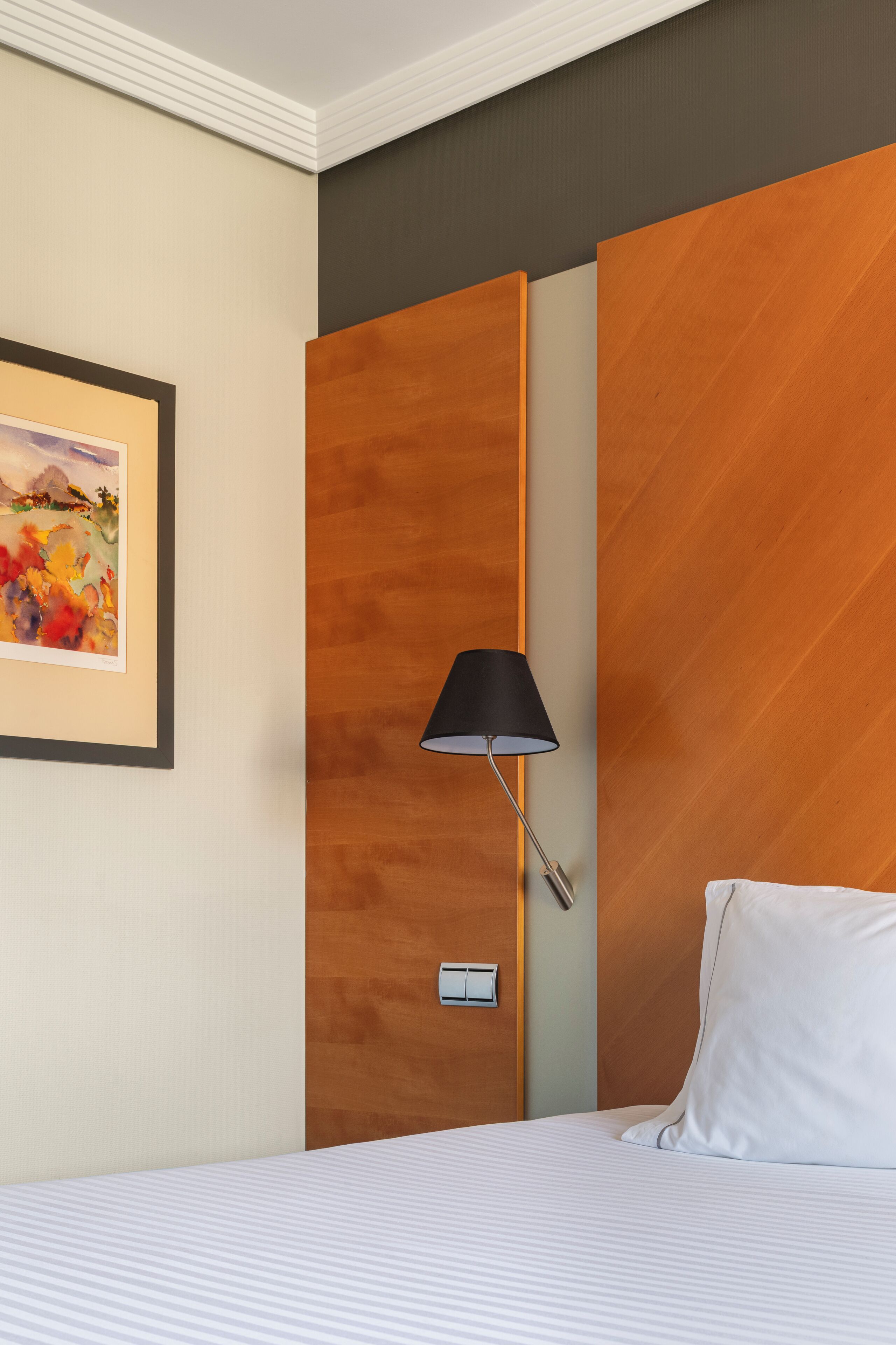 A hotel room with a bed, a framed picture on the wall, a bedside lamp, and a wooden headboard.