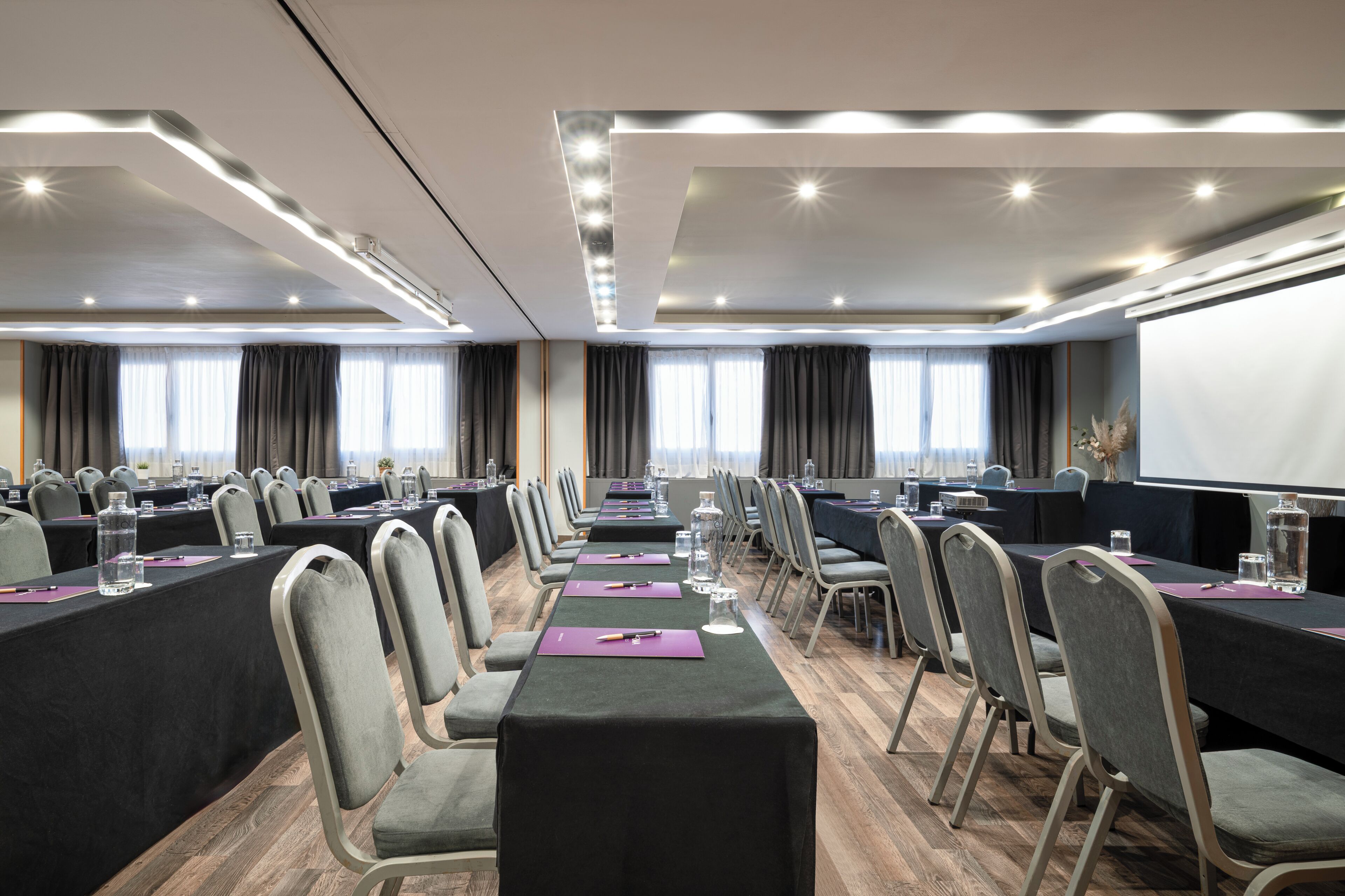 Un salón de conferencias con filas de mesas, sillas y una pantalla de proyector.