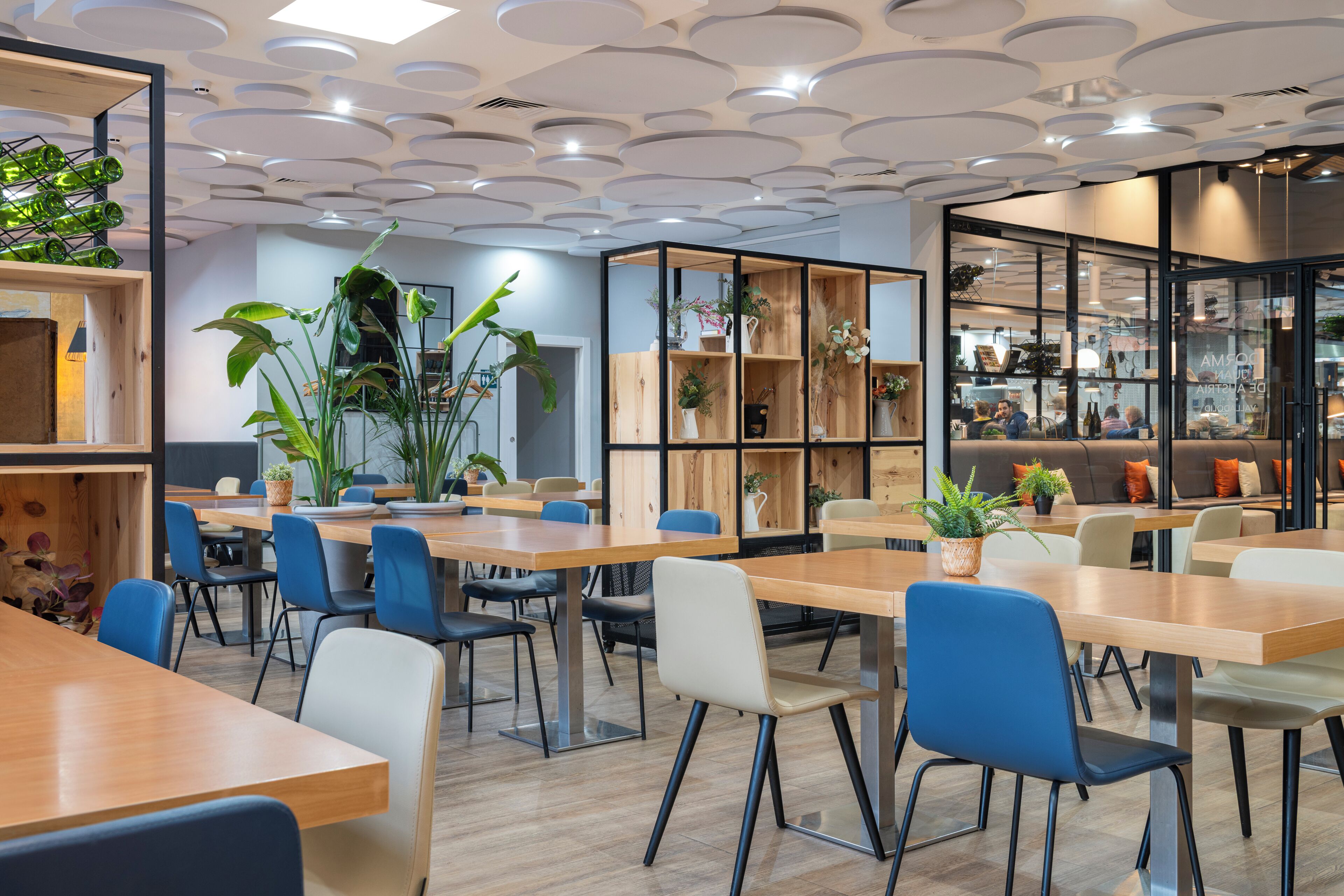 A modern dining area with wooden tables and chairs, shelves with plants, and a view into a kitchen area.
