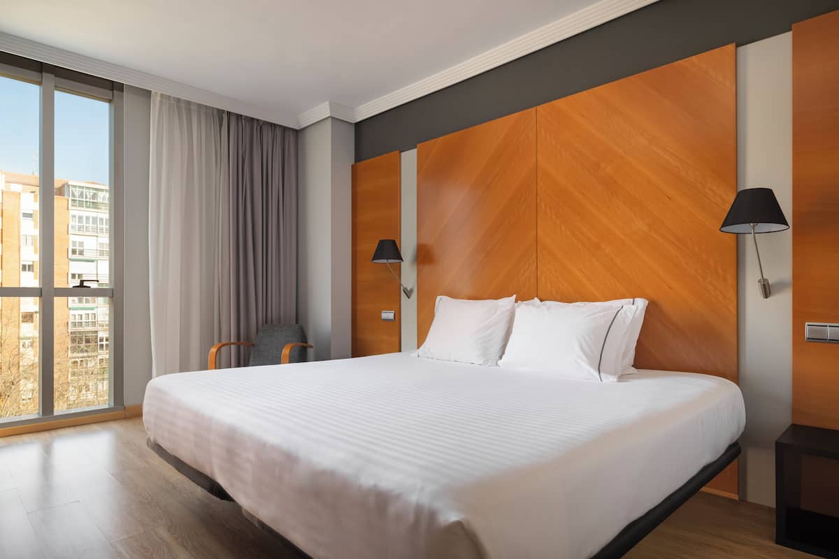 A hotel room with a large bed, a view of buildings, a chair, and a lamp.