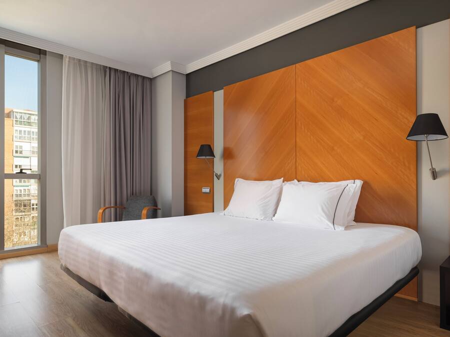 A hotel room with a large bed, a view of buildings, a chair, and a lamp.