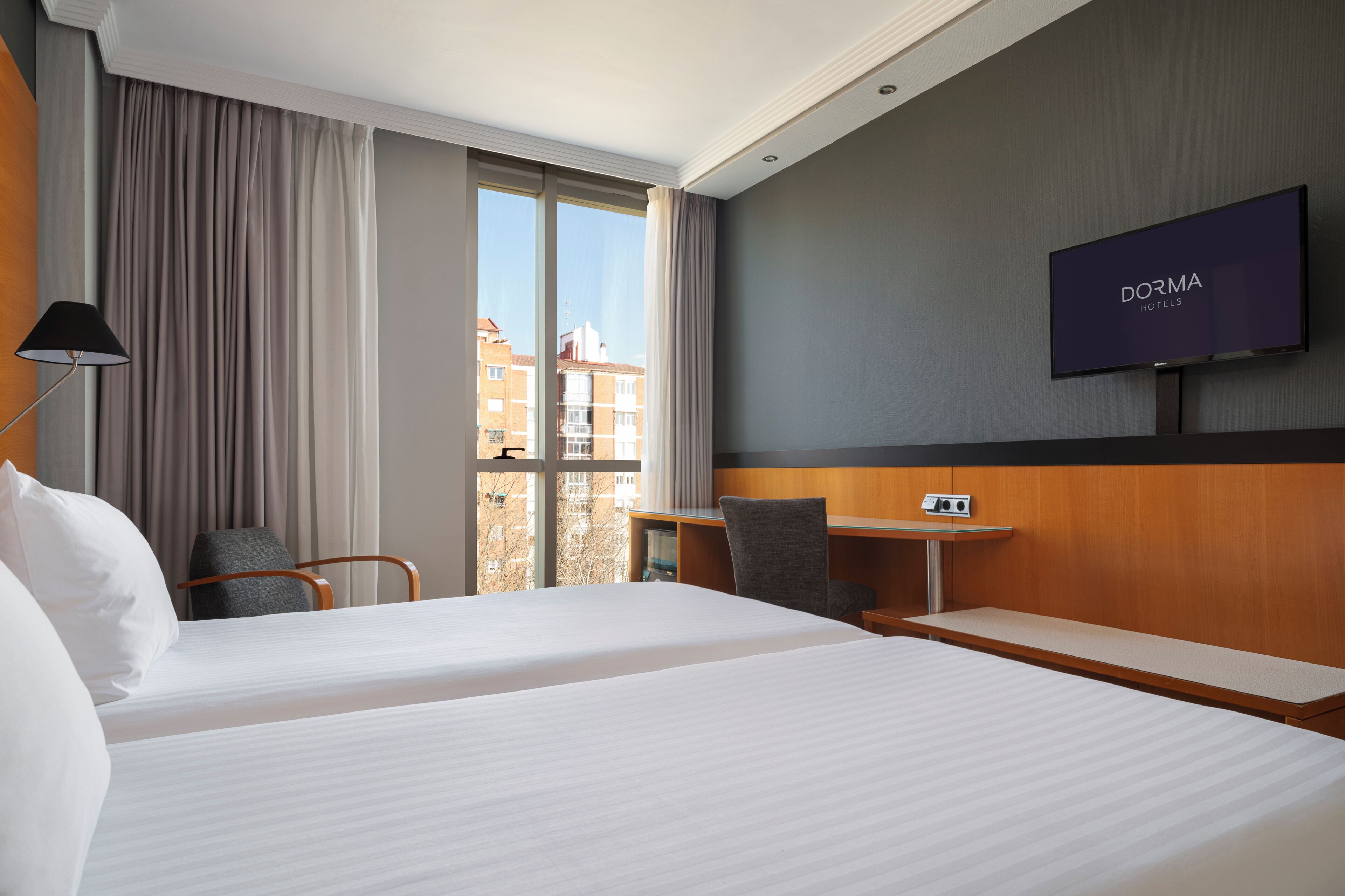 A hotel room with two beds, a TV, a desk, and a view of buildings outside.