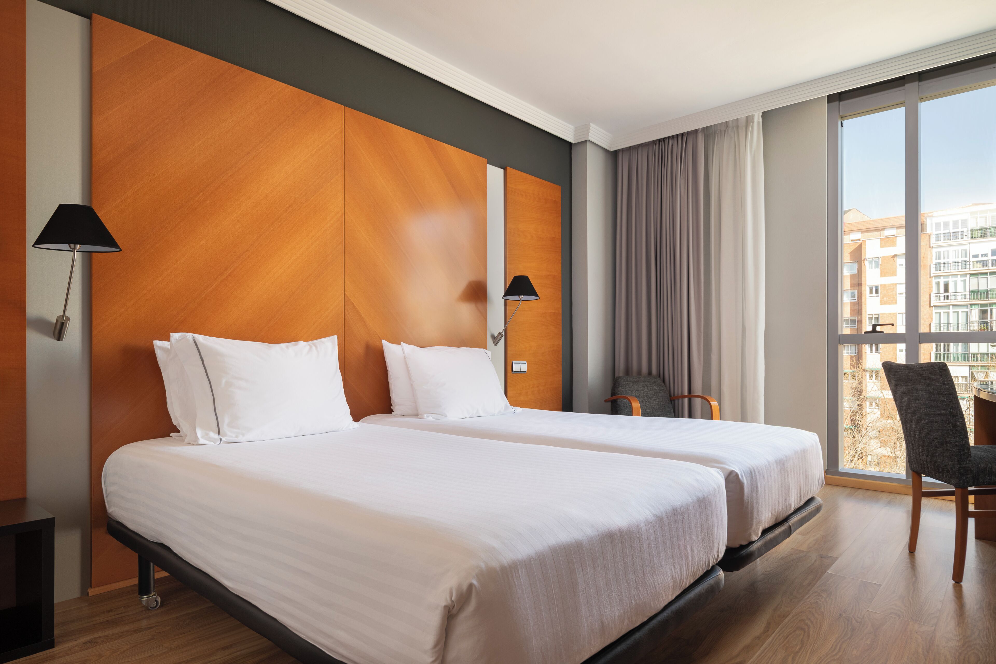 A hotel room with two beds, a view of buildings, and a chair.