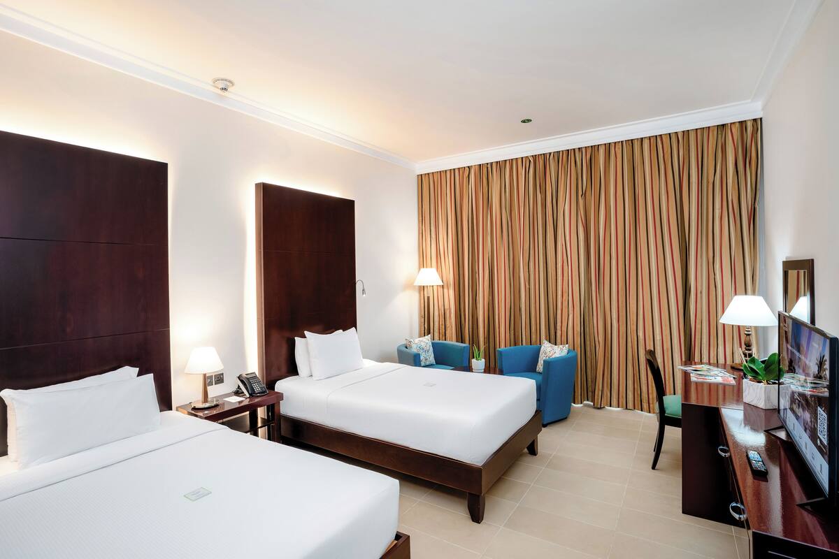 A hotel room with two beds, a desk with a TV, and a large window with curtains.