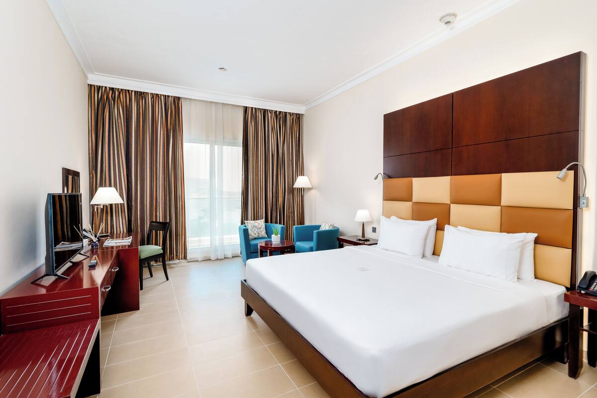 A hotel room with a large bed, a desk with a lamp, a chair, and a TV.