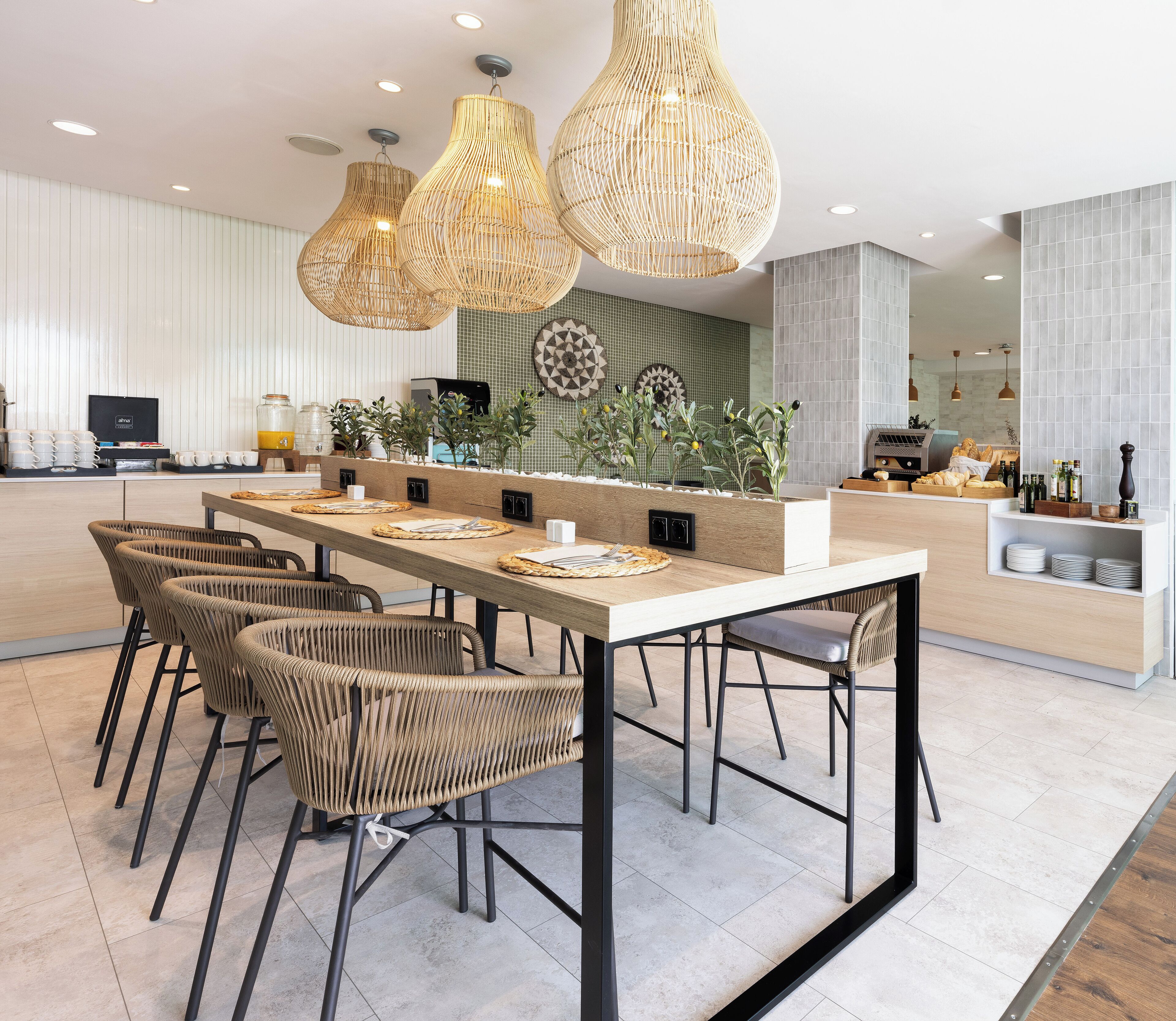 Un espace de restauration moderne avec une longue table, des chaises en osier et des luminaires suspendus.