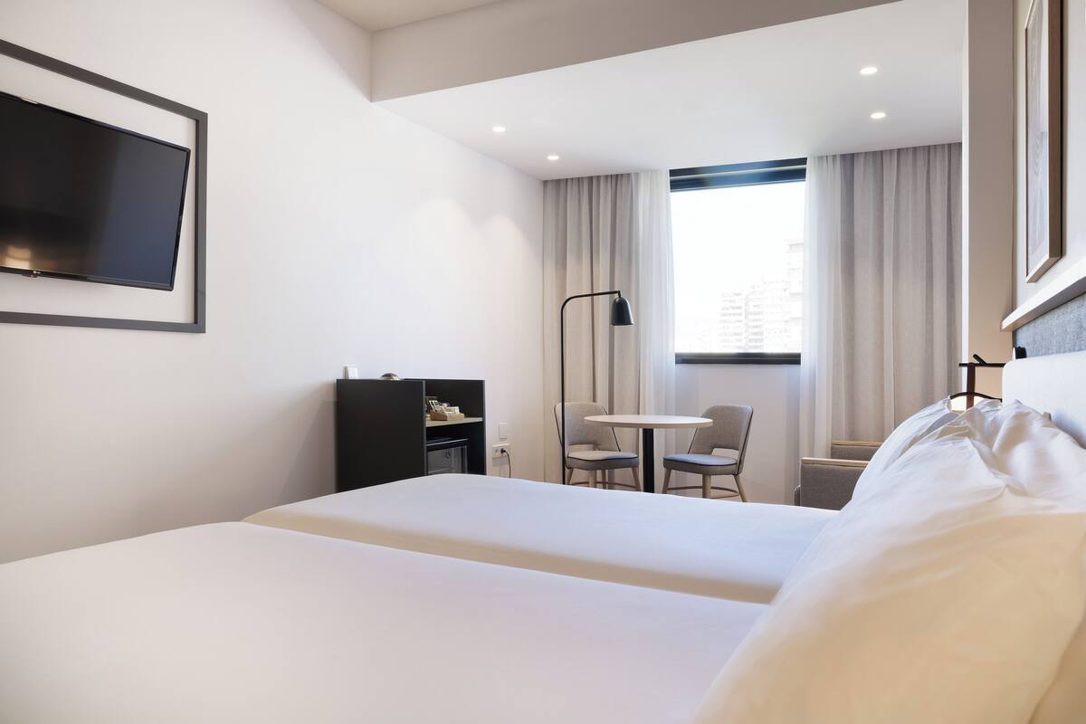Hotellihuone, jossa on kaksi sänkyä, työpöytä, tuoli, televisio ja ikkuna, jossa on verhot.
