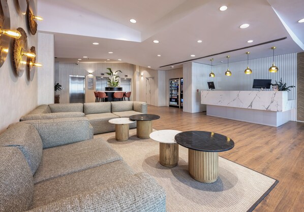 A modern lobby with a sectional sofa, round tables, a reception desk, and a vending machine.