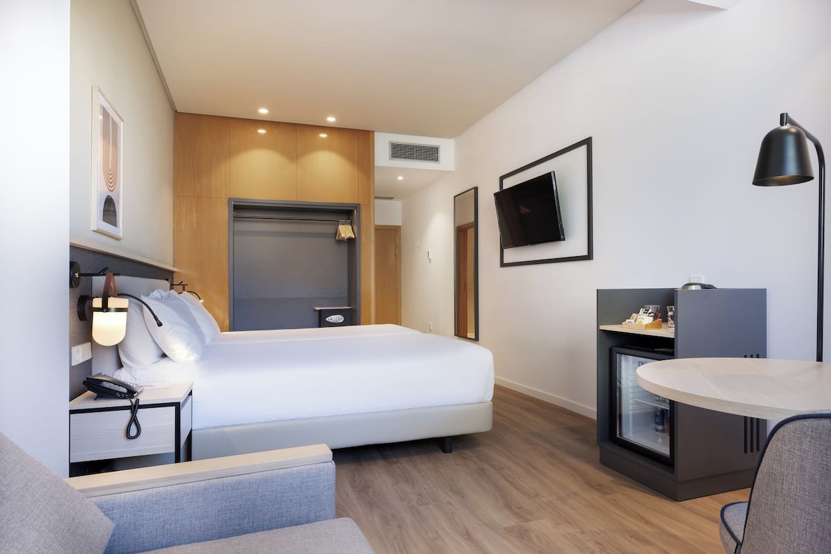 Moderni hotellihuone, jossa on sänky, yöpöytä, lamppu, televisio ja pieni keittonurkkaus.