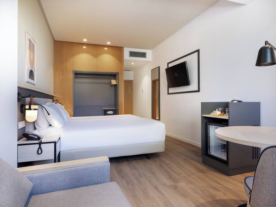 Una habitació d'hotel moderna amb llit, taula de nit, làmpada, televisió i una petita zona de cuina.