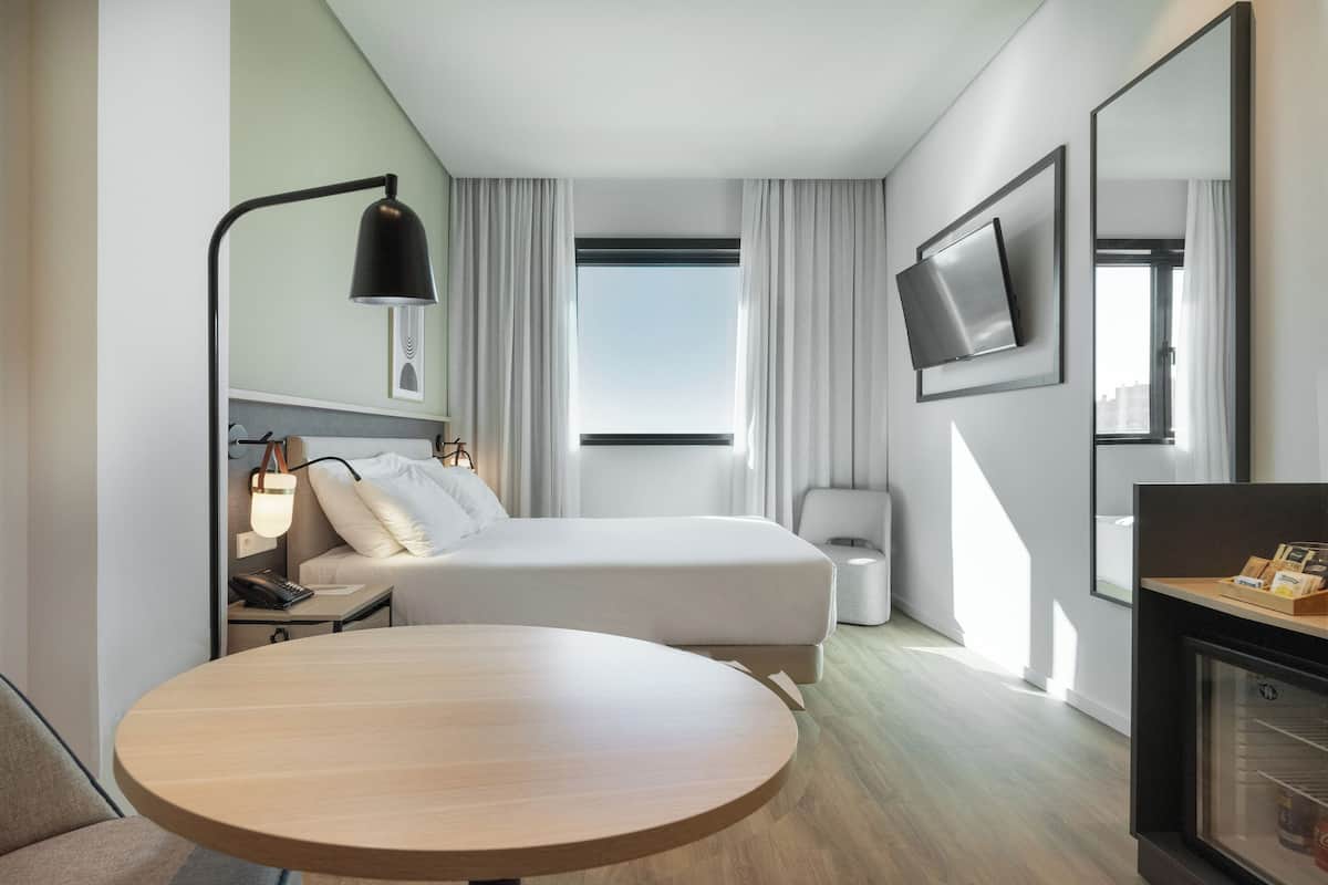 Moderni hotellihuone, jossa on sänky, yöpöytä, lamppu, televisio ja pyöreä pöytä.