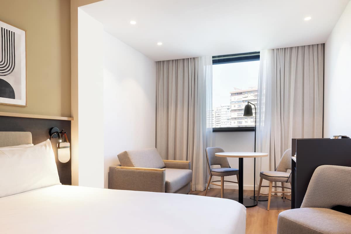 Moderni hotellihuone, jossa on suuri sänky, työpöytä, kaksi tuolia ja ikkunasta avautuva näkymä kaupunkiin.