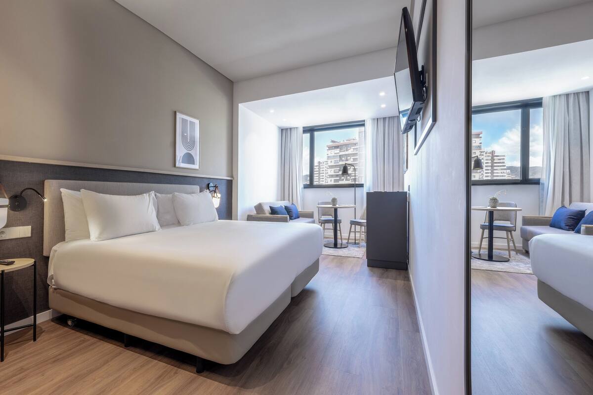 Moderni hotellihuone, jossa on suuri sänky, työpöytä ja oleskelualue, josta on näkymä kaupunkiin.