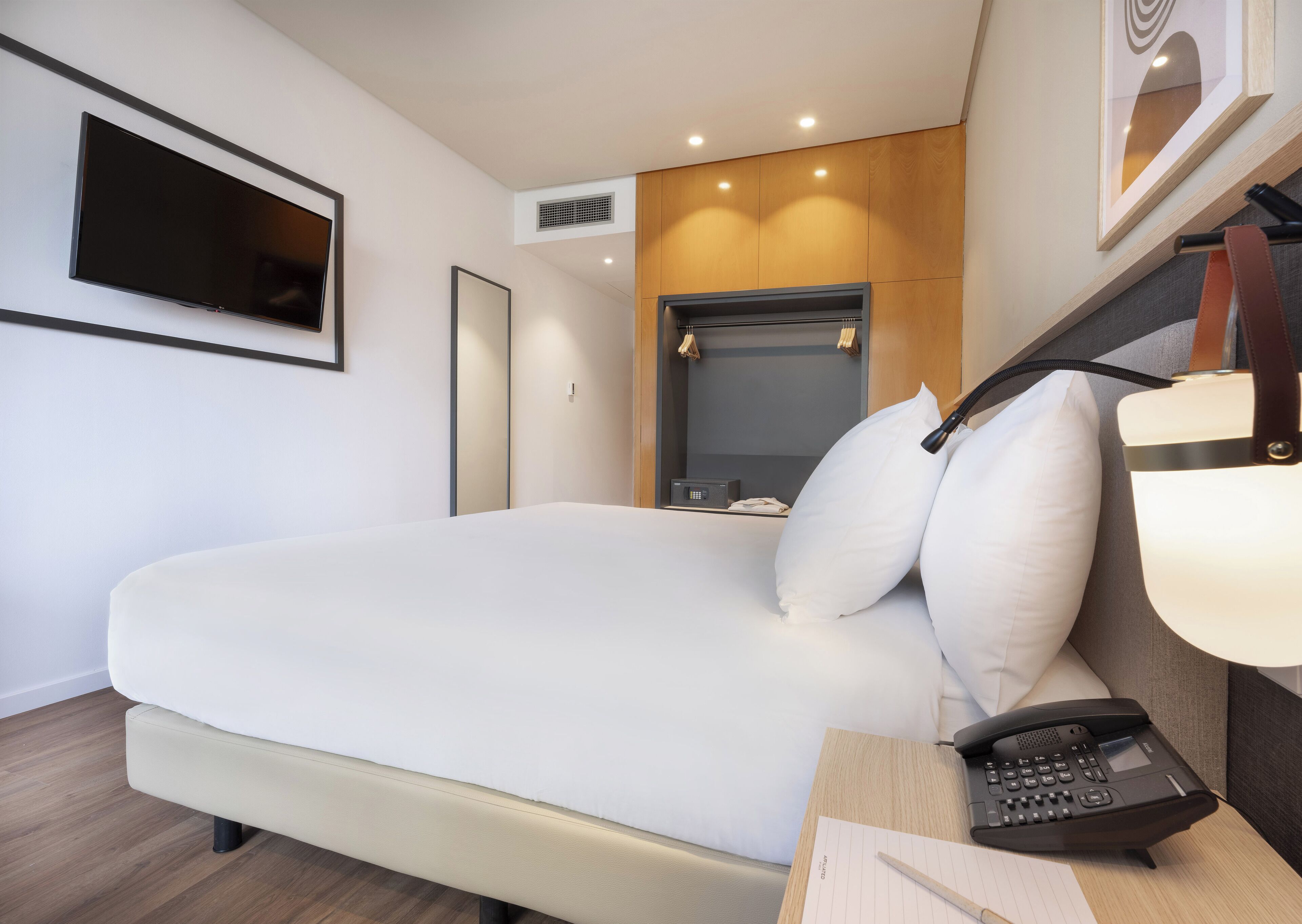 Ein modernes Hotelzimmer mit einem großen Bett, einem Nachttischtelefon, einem Schreibtisch mit Lampe und einem an der Wand befestigten Fernseher.