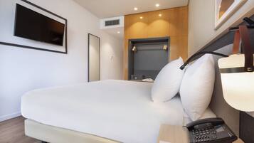 Ein modernes Hotelzimmer mit einem großen Bett, einem Nachttischtelefon, einem Schreibtisch mit Lampe und einem an der Wand befestigten Fernseher.