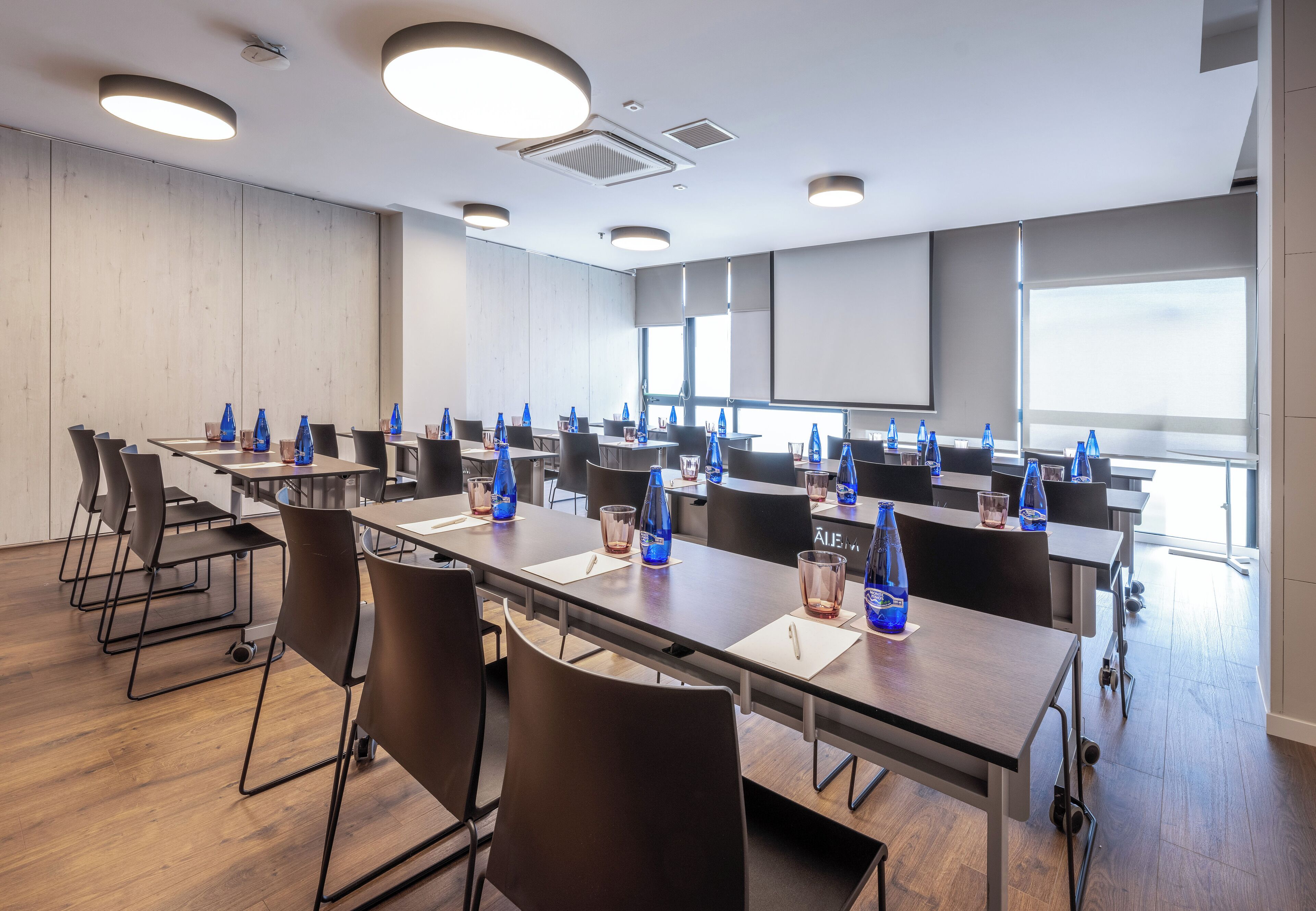 A conference room with multiple tables and chairs, equipped with water bottles and glasses, and large windows providing natural light.