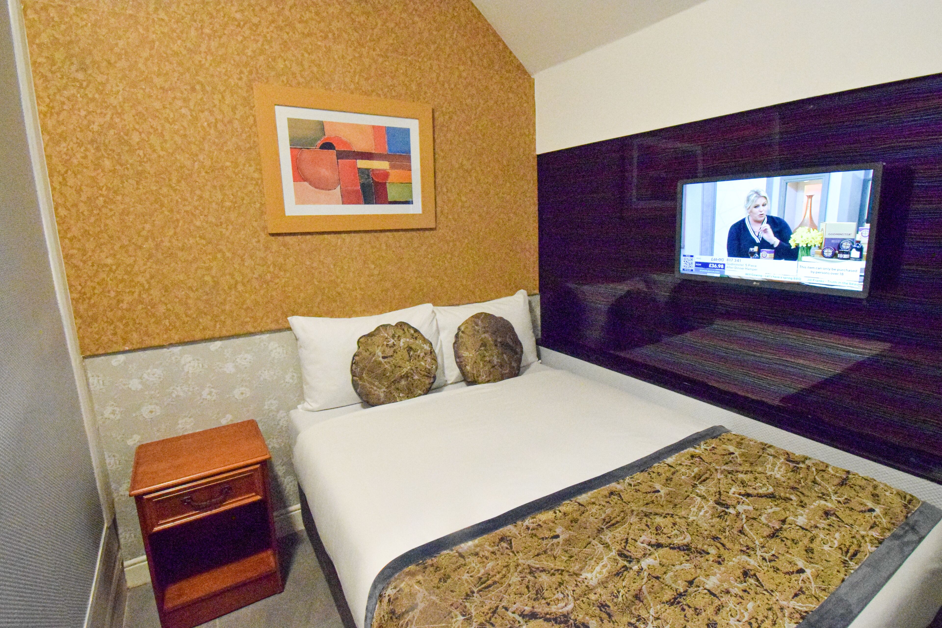 A hotel room with a bed, a television on the wall, a small wooden nightstand, and a framed picture on the wall.