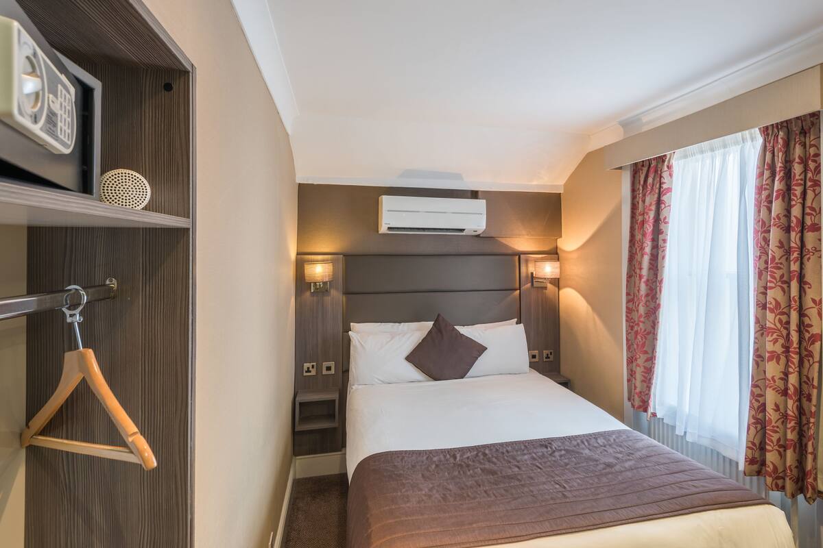 Camera d'albergo con un letto, lampade sui comodini, un condizionatore d'aria, un appendiabiti in legno e uno scaffale con un oggetto decorativo.