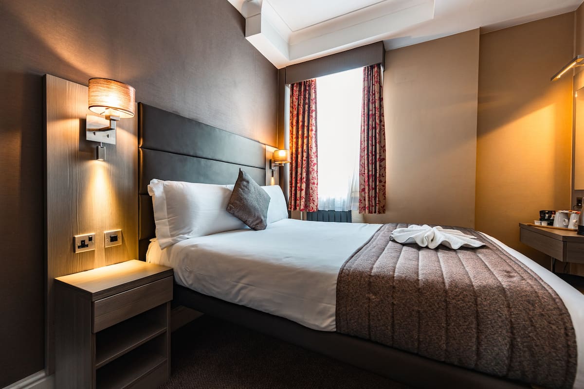 Una camera d'albergo con un letto grande, un comodino, una lampada a parete, una finestra con tende e un tavolino con una macchina per il caffè.
