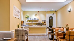 A dining area with a buffet setup, including a counter with various food items and a selection of plates and bowls. There are several dining tables with chairs, and a wall-mounted light fixture above the counter. - Brunel Hotel (London)