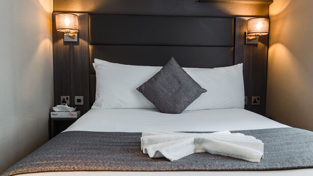 A neatly made bed with a grey pillow, a white pillow, and a folded white towel on a grey blanket. There are two wall-mounted lamps on either side of the bed. - Brunel Hotel (London)
