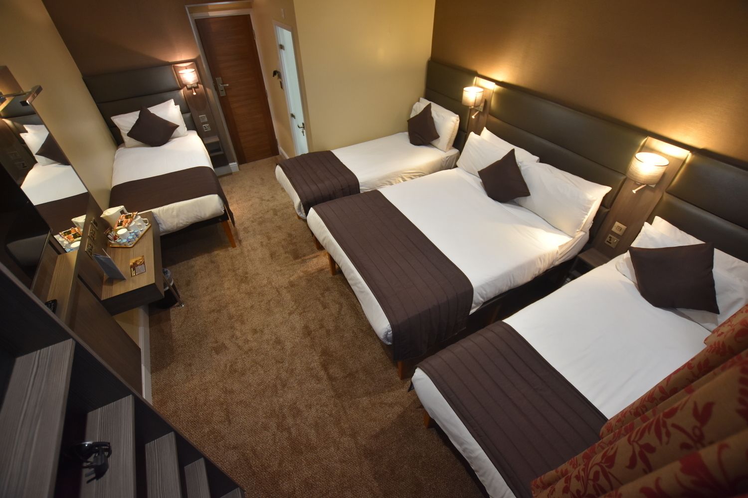 Una habitación de hotel con cuatro camas dispuestas en fila, cada una con cabecera y lámparas de noche.  A la izquierda de la habitación hay una mesita con una lámpara y un reloj.