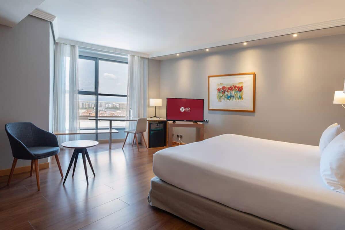 A hotel room with a bed, a desk with a computer, a chair, a TV, and a window with a city view.