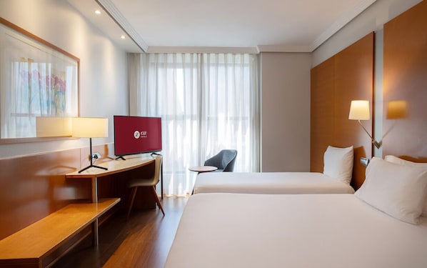 Habitación de hotel con escritorio, dos camas, televisor y ventana con cortinas traslúcidas.