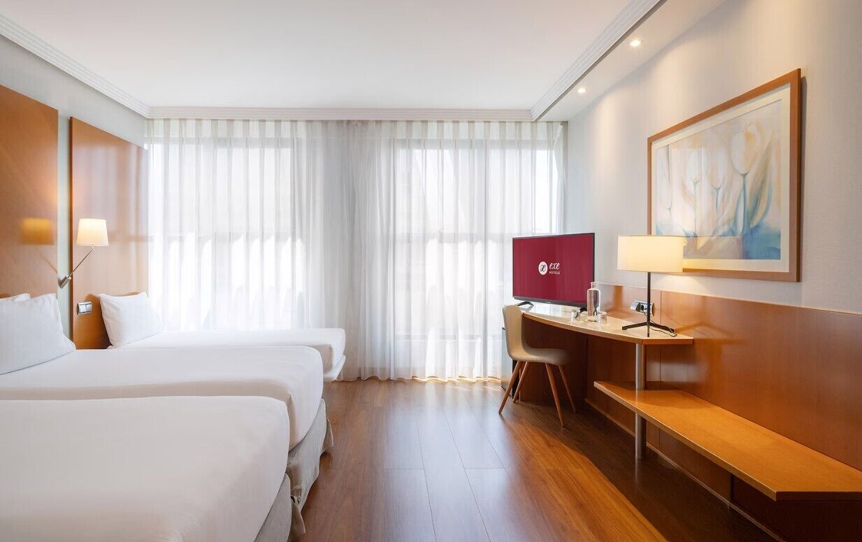 Et hotelværelse med to senge, et skrivebord med en computer og et stort vindue med lette gardiner.