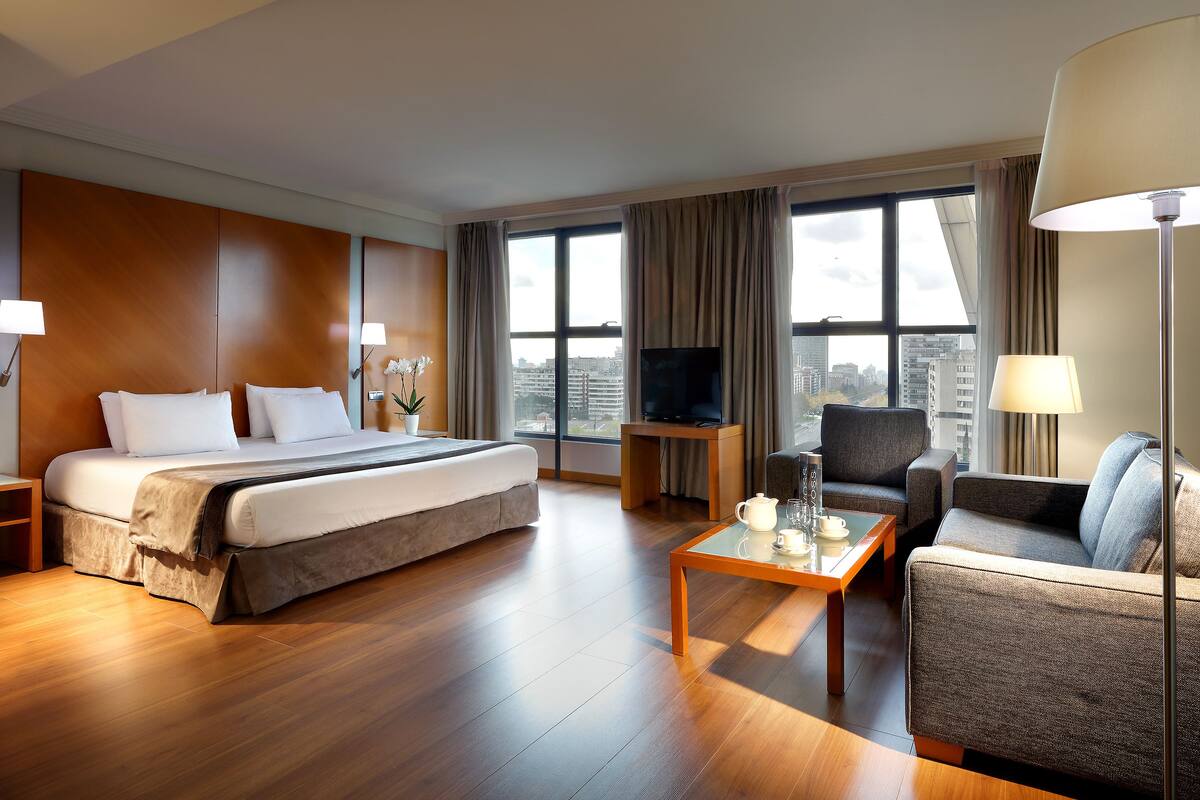A hotel room with a large bed, a sofa, a coffee table, a TV, and a city view.