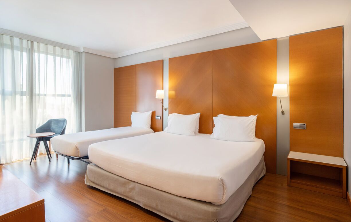 Una camera d'albergo moderna con due letti, un tavolino, una sedia e un'ampia finestra con tende.