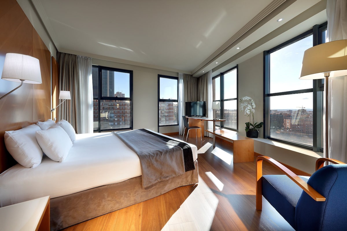 A modern hotel room with a large bed, a desk, a chair, and large windows offering city views.