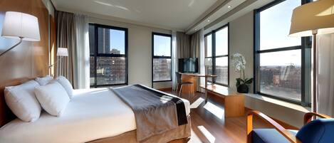 Habitación de hotel moderna con una cama grande, un escritorio, una silla y ventanas amplias que ofrecen vistas a la ciudad.