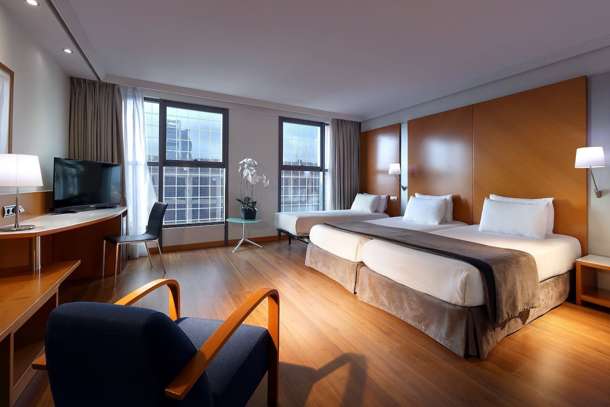 A hotel room with a bed, desk, chair, TV, and large windows.