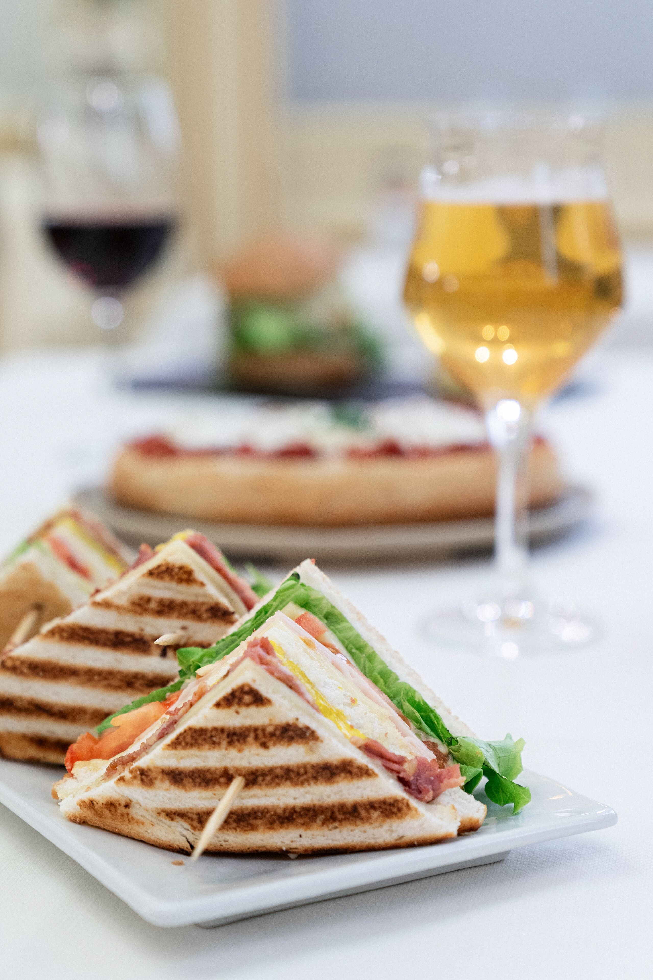 A plate of triangular sandwiches with visible layers of ingredients, a glass of white wine, and a blurred dessert in the background.