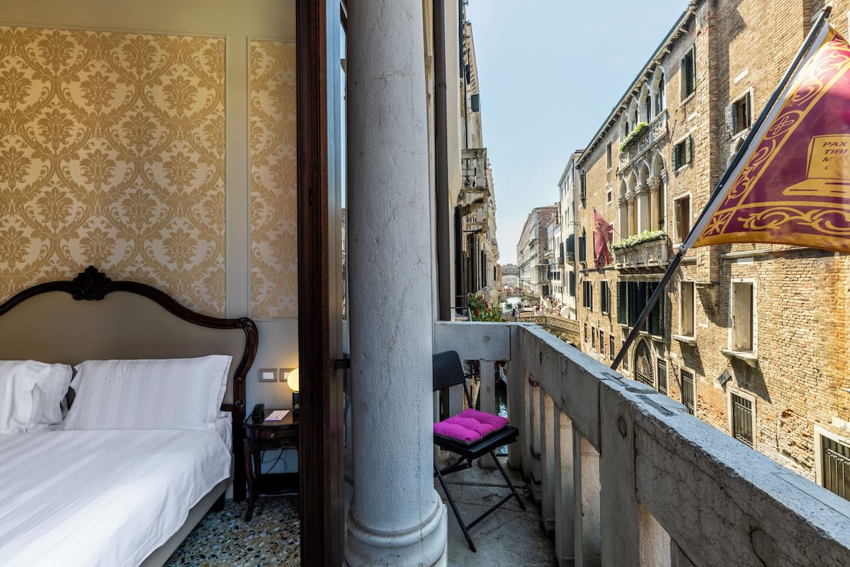 A hotel room with a bed, a small table with a lamp, a chair, and a balcony overlooking a street with buildings and a flag.