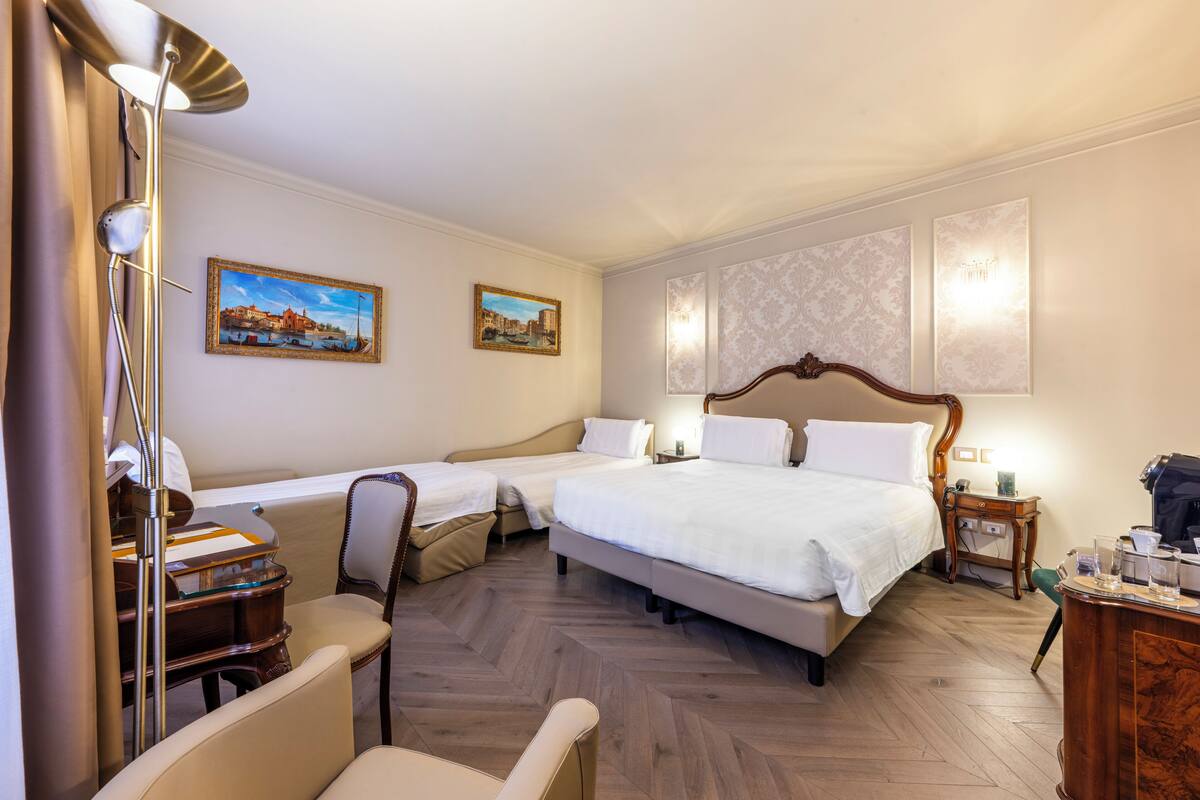 A hotel room with two single beds, a large bed, a desk with a chair, a lamp, and two paintings on the wall.