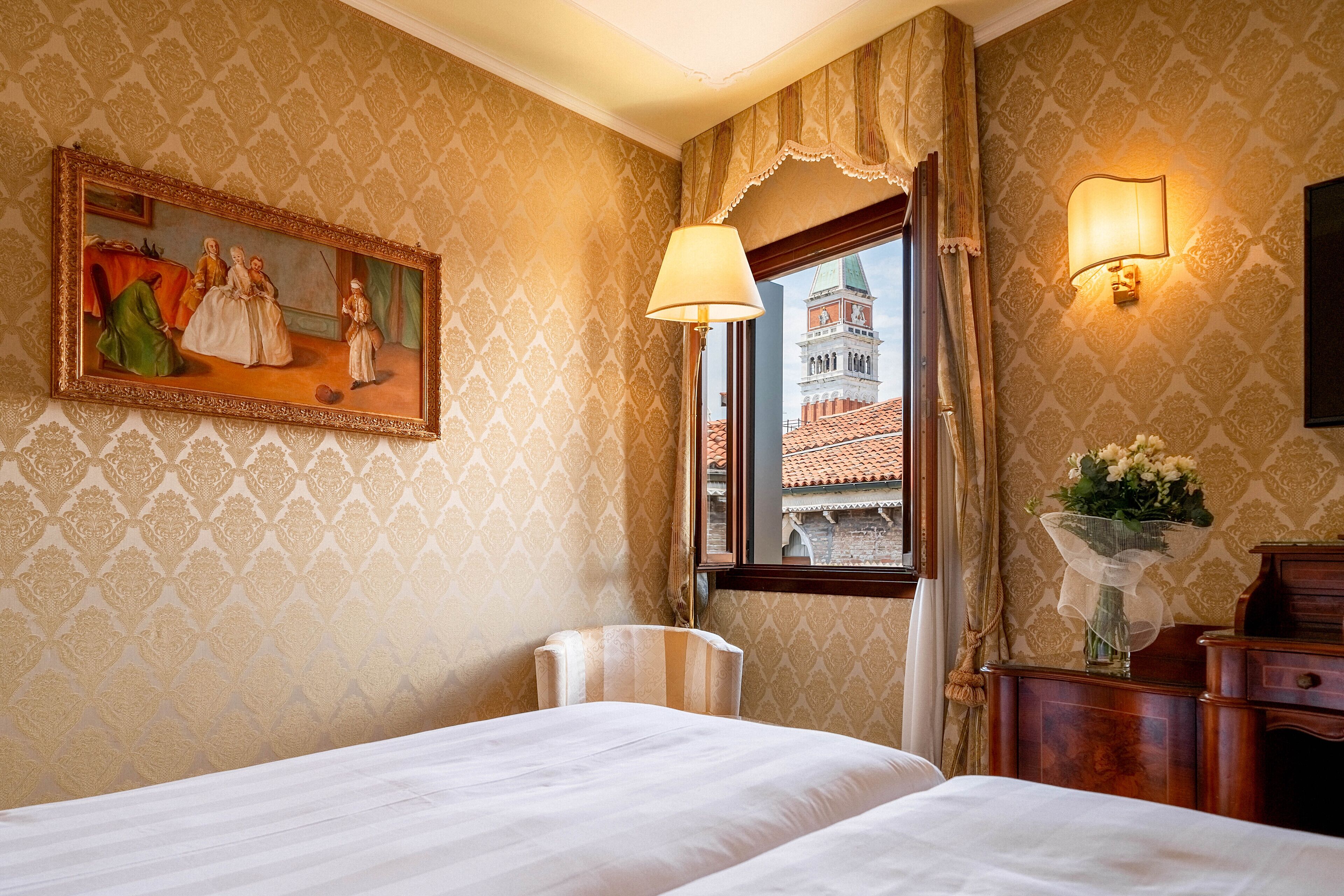 Camera d'albergo con un letto, una sedia, un quadro, una lampada, un vaso con fiori e vista su un edificio attraverso una finestra.