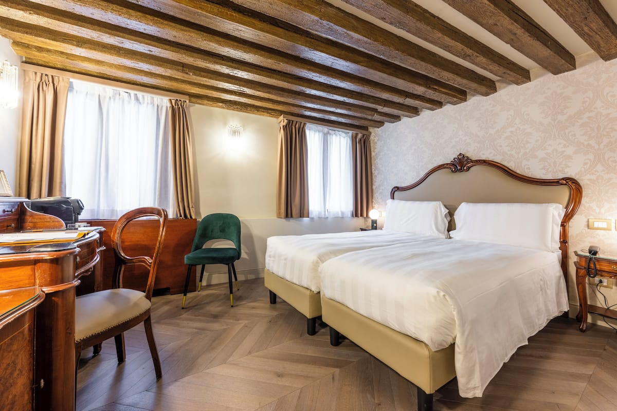 A hotel room with a wooden ceiling, two beds, a desk with a chair, and a small table with a lamp.