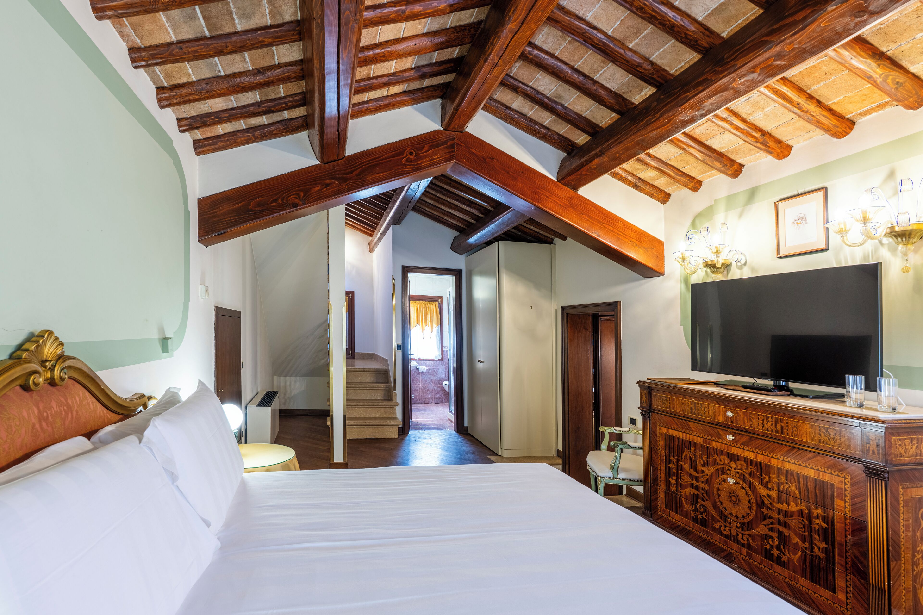 Habitación de hotel con una cama grande, vigas de techo de madera, un televisor sobre un mueble de madera y una escalera que conduce a otra habitación.