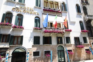 A hotel with balconies adorned with flowers, European flags, and a sign reading "HOTEL COLOMBINA".