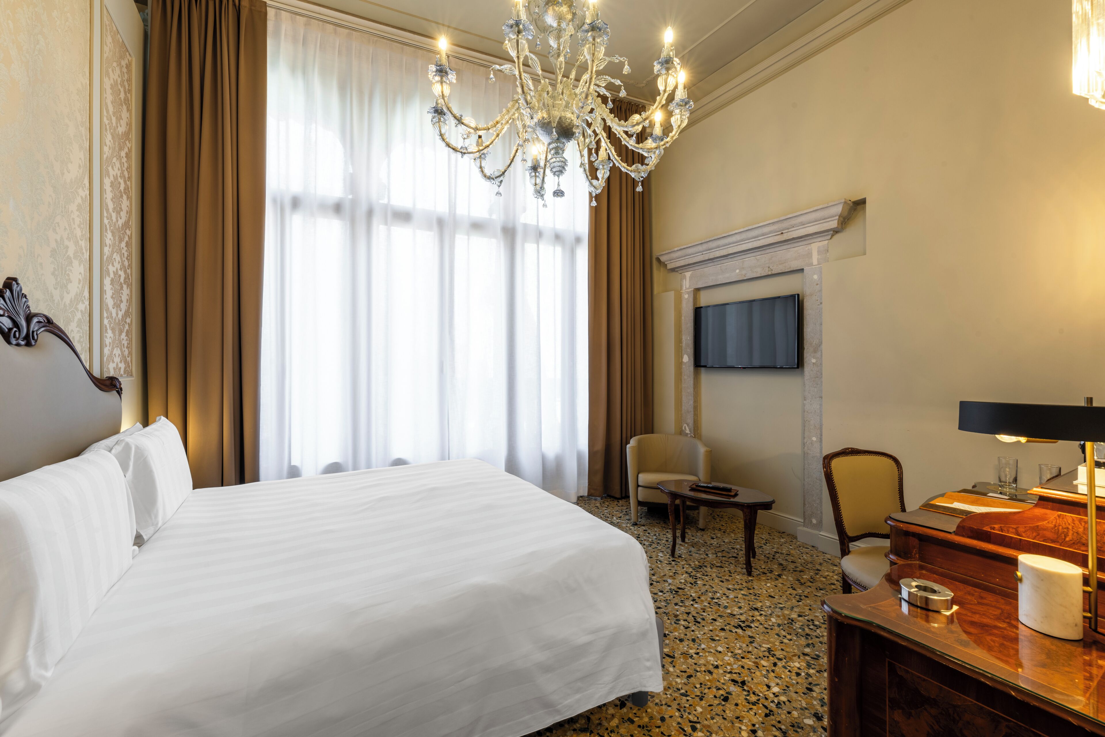 A hotel room with a large bed, a chandelier, a fireplace, and a desk with a lamp.