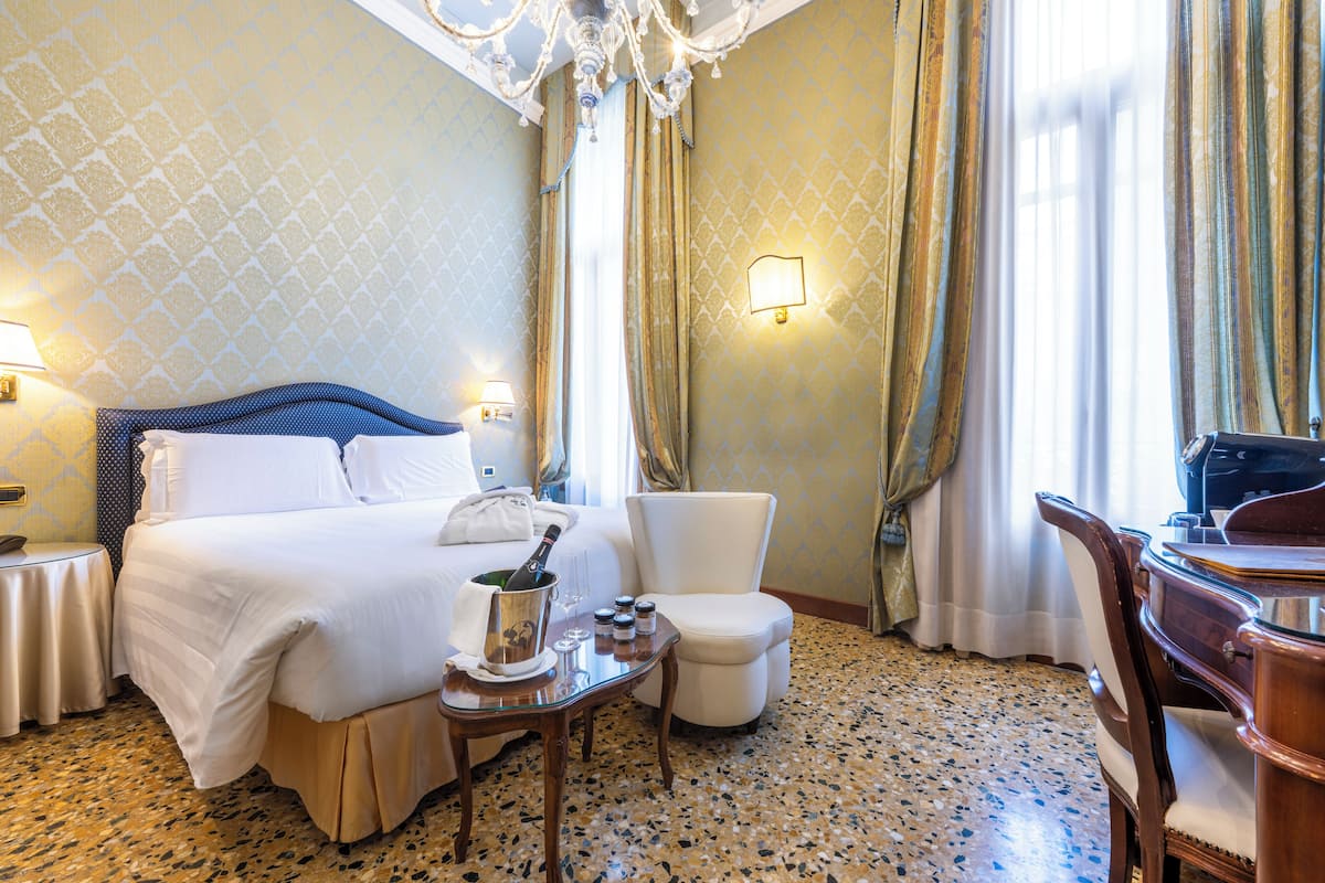A hotel room with a bed, a chair, a desk, a chandelier, and a wall-mounted lamp.