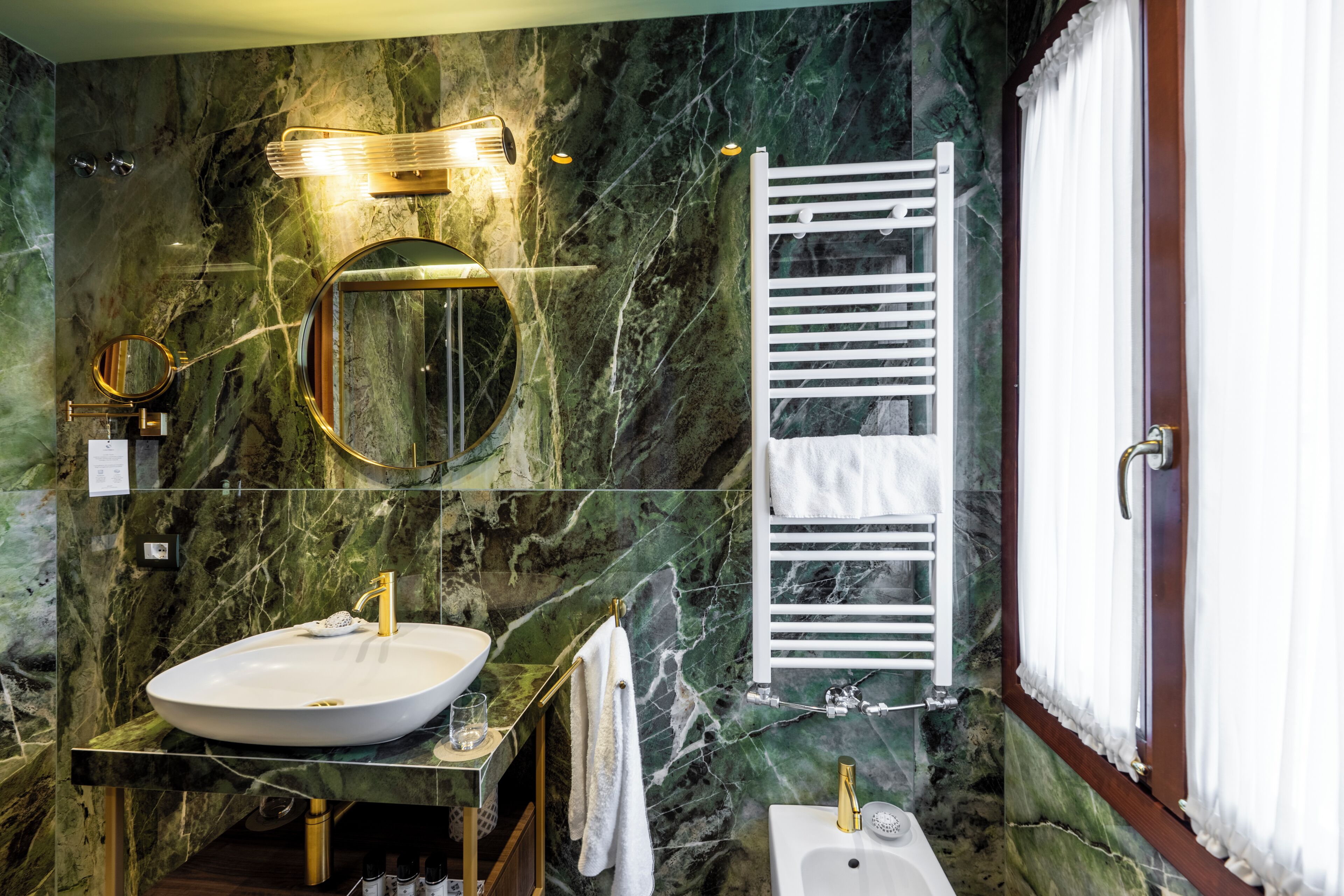 Un bagno con parete in marmo, uno specchio tondo, un lavabo, un porta asciugamani e una finestra con tende.