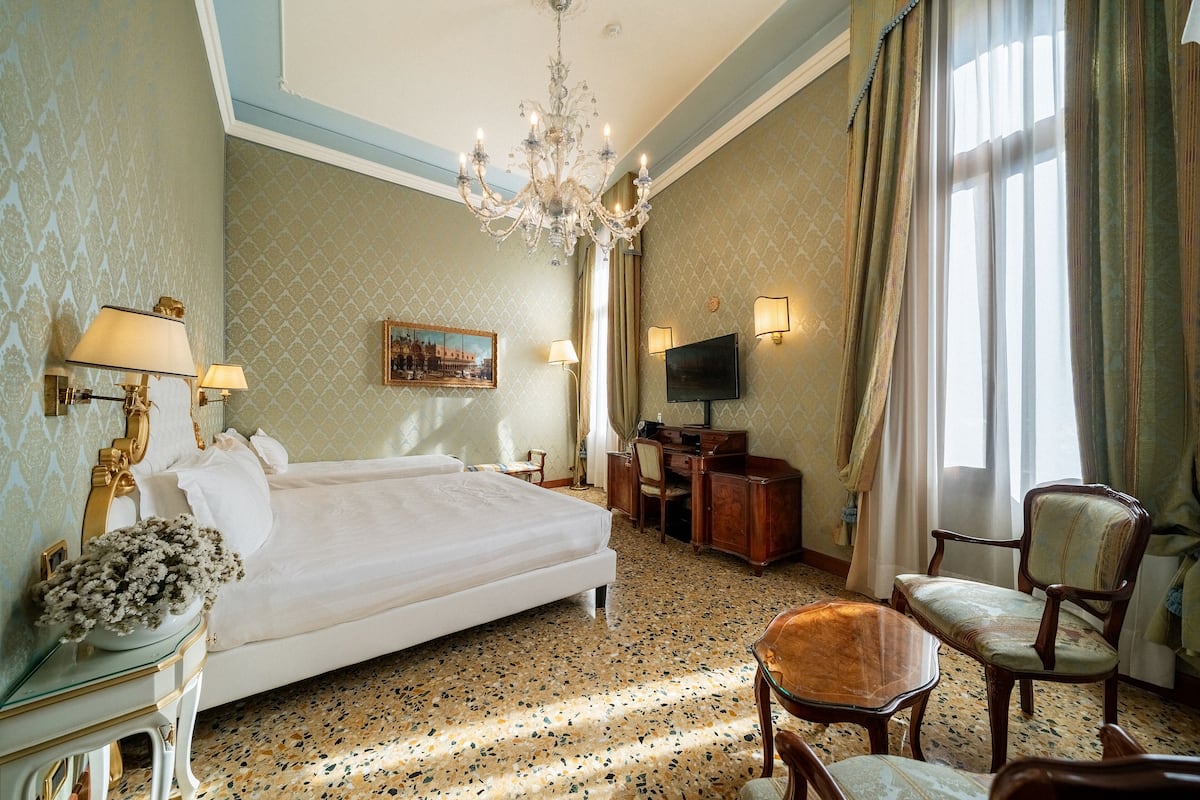 A hotel room with a large bed, a desk with a TV, a chandelier, and a window with curtains.