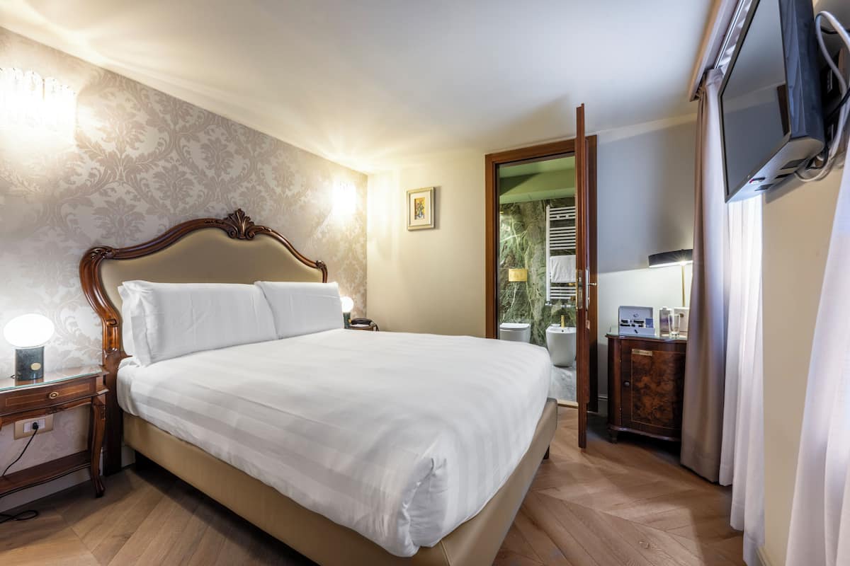 A neatly made bed in a hotel room with a wooden headboard, a bedside table with a lamp, and a television mounted on the wall.